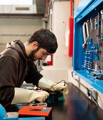 Young engineer testing electronic device in laboratory, wearing protective gloves and holding multimeter, concentrating on technical measurement